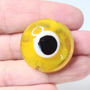 May include: A yellow glass bead with a blue and white evil eye design. The bead is round and has a hole in the center for stringing.