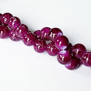 May include: A strand of round, purple glass beads with white swirls. The beads are strung on a clear thread.