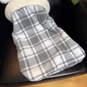 May include: A grey and white plaid fleece blanket with a cream-coloured, fluffy, sherpa-lined opening. The blanket is folded and placed on a black leather chair. The blanket is designed to keep you warm.