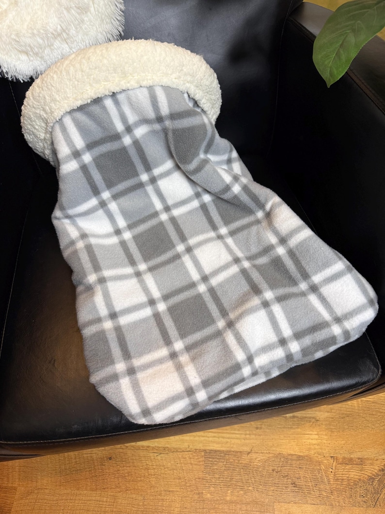 May include: A grey and white plaid fleece blanket with a cream-coloured, fluffy, sherpa-lined opening. The blanket is folded and placed on a black leather chair. The blanket is designed to keep you warm.