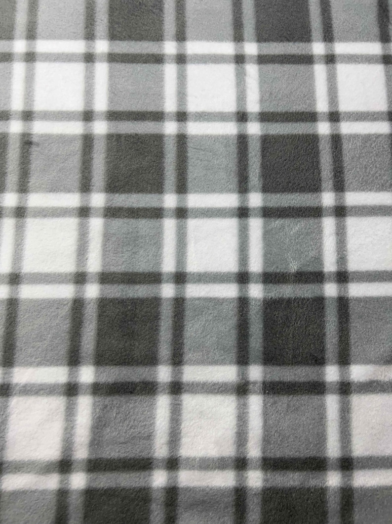 May include: A close-up of a soft, grey and white plaid fleece fabric. The pattern consists of intersecting lines forming squares and rectangles in varying shades of grey and white. The fabric has a fuzzy texture.