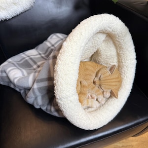 May include: A ginger tabby cat sleeping inside a cream-coloured pet bed with a fluffy white rim. The bed is on a black leather chair, with a grey and white plaid blanket partially visible. The cat is curled up and asleep.