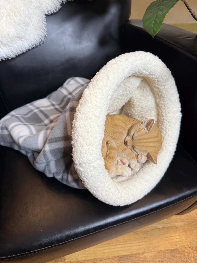 May include: A ginger tabby cat sleeping inside a cream-coloured pet bed with a fluffy white rim. The bed is on a black leather chair, with a grey and white plaid blanket partially visible. The cat is curled up and asleep.