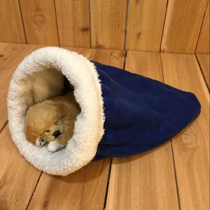 May include: A cozy blue pet bed with a white, fluffy interior. A small, tan-colored puppy is nestled inside, appearing to be sleeping. The bed is made of soft materials, providing a comfortable space for a small dog or cat.