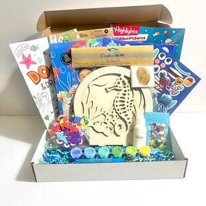 May include: A craft kit in a white box containing a wooden seahorse layer art project, a Highlights Hidden Pictures book, a glue bottle, small paint pots in various colors, a bag of colorful sequins, foam sea creature shapes, and blue paper filler.