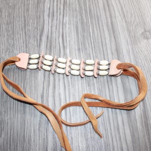 May include: A choker necklace featuring three horizontal rows of white cylindrical beads separated by small silver-toned metal spacers. The beaded section is attached to two light pink leather tabs connected to long, tan suede leather ties.