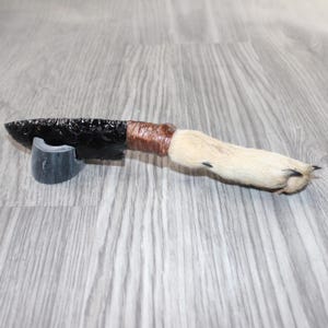 May include: A handcrafted obsidian knife with a fur handle and a brown wrapped grip. The blade is black and rests on a gray stand. The handle is made of light-colored fur with black claws.