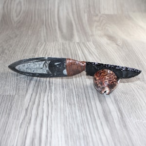 May include: A handcrafted obsidian knife with a fossilized handle and a wrapped brown leather grip. A small, patterned seashell rests near the blade. The knife is displayed on a wooden surface.