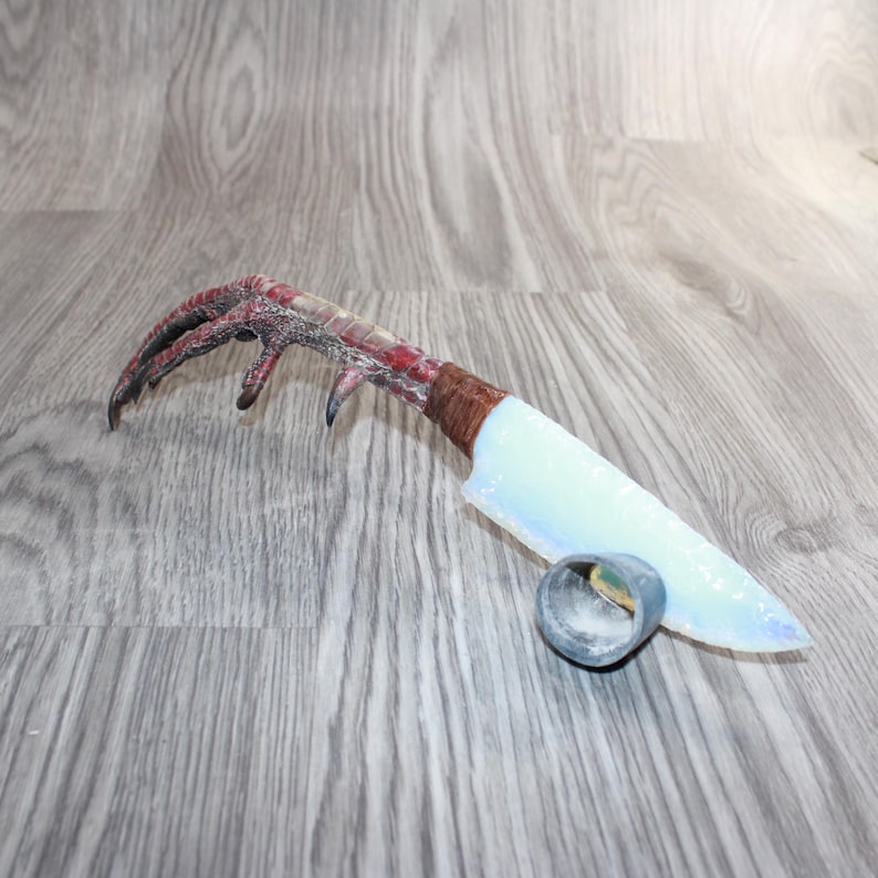 May include: A distinctive knife featuring an opalescent blade and a handle designed to mimic a bird's foot. The handle is wrapped in brown material, with a small, silver-toned ring at the blade's base. A unique decorative item.