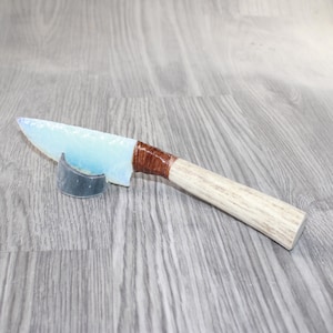 May include: A handcrafted knife with a light blue, translucent blade. The handle is made of light-colored wood and wrapped with brown material. The knife rests on a small, dark gray stand.