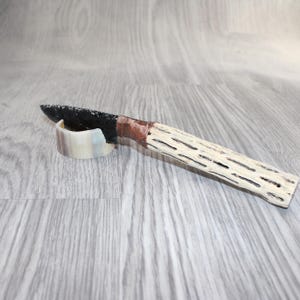 May include: A handcrafted knife with a black blade and a white, textured handle. The handle is made from a natural material, and the blade is supported by a small, curved stand. The handle is 6 inches long.