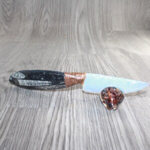 May include: A knife with a pale blue translucent blade and a dark grey handle featuring fossilized patterns. The handle is connected to the blade with brown cord wrapping. The knife rests on a small, brown, spotted, oval-shaped seashell.