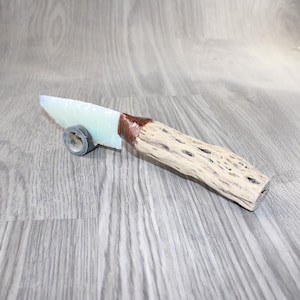 May include: A handcrafted knife with a light green blade and a wooden handle. The blade is held in place with a brown wrapping. The handle is made of light-colored wood with a textured surface. The knife rests on a small, circular, gray stand.