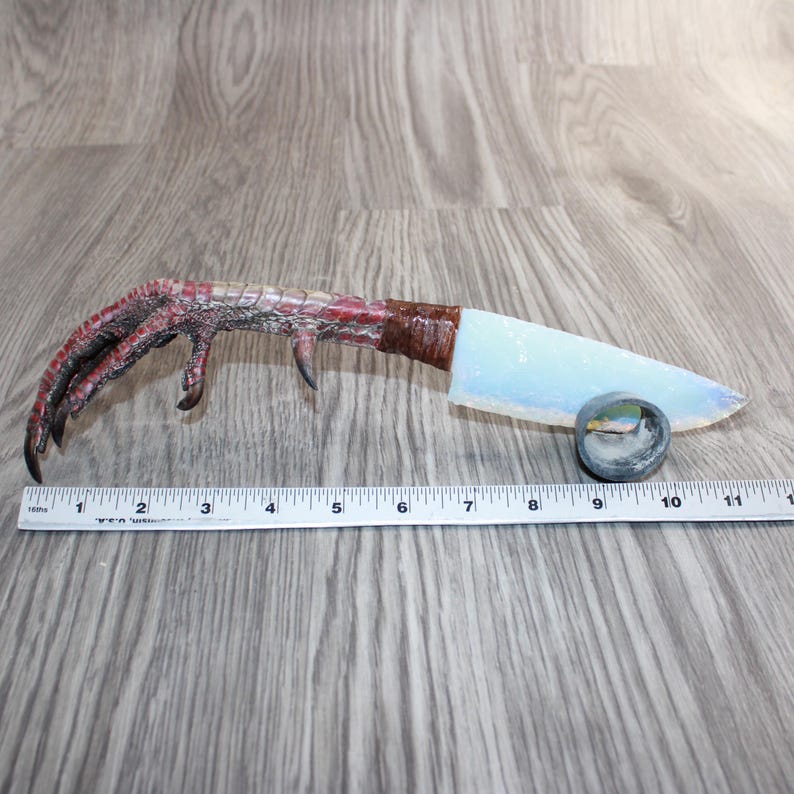 May include: A novelty knife featuring a bird claw handle and a light blue blade. The handle is a detailed bird claw in red and brown hues. The blade measures approximately 10 cm. A small, round object is positioned near the blade.