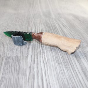 May include: A handcrafted knife with a green, textured blade and a light brown, fur-covered handle. The handle is wrapped with brown material near the blade. The knife rests on a gray surface.