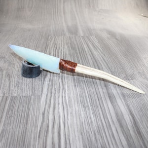 May include: A handmade spear point with a pale blue, translucent blade. The blade is attached to a light-colored, curved shaft with a brown, wrapped handle. The spear point rests on a small, black, cylindrical stand.