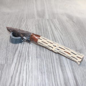 May include: A handcrafted knife with a dark stone blade and a handle made from a light-colored, textured material. The handle is wrapped with a brown material near the blade. The knife rests on a small, gray stand.