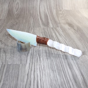 May include: A decorative knife with a light blue blade and a white handle. The handle has a textured, ridged design. The blade is supported by a small, curved, gray stand. The handle is wrapped in brown material.