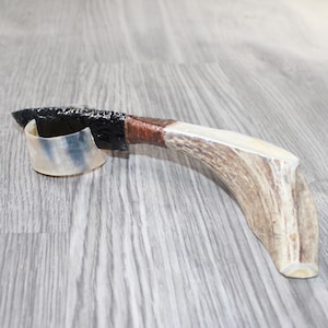May include: A handcrafted obsidian blade knife with a handle made from antler and wrapped in brown leather. The knife features a curved antler handle and a small, curved, translucent piece at the base of the blade.