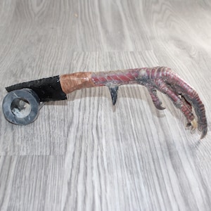 May include: A smoking pipe featuring a realistic bird claw as the stem, connected to a black obsidian-like blade and a cylindrical dark grey stone bowl. The claw displays textured reddish-brown scales and sharp, curved talons at the end.