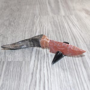 May include: A knife with a reddish-brown stone blade and a dark, curved handle made of animal horn. The handle and blade are joined by a section of wrapped brown cord. The knife is resting on a small, black, conical display stand.