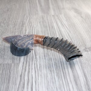 May include: A tool featuring a sharp, grey, knapped stone blade attached to a curved, dark, ridged animal horn handle. The connection point between the stone blade and the horn handle is wrapped in thin, light brown cordage for secure attachment.