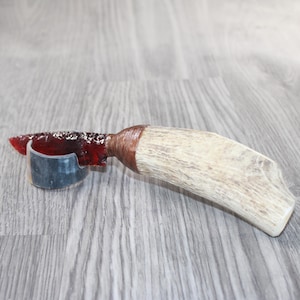 May include: A handmade knife with a red, translucent blade and a light-colored antler handle. The blade is held in a dark gray holder. The handle is wrapped in brown material.
