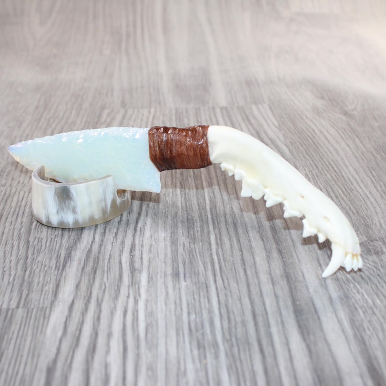 May include: A handcrafted knife with a light blue blade and a brown wrapped handle, paired with a bone-like jaw. The knife rests on a decorative, curved holder. The design evokes a primitive, natural aesthetic.