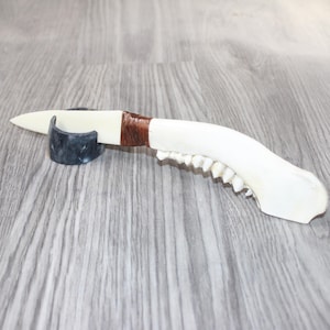 May include: A handcrafted knife with a bone handle and a blade. The handle is wrapped with brown material. The knife rests in a dark gray holder. The handle has a row of teeth.