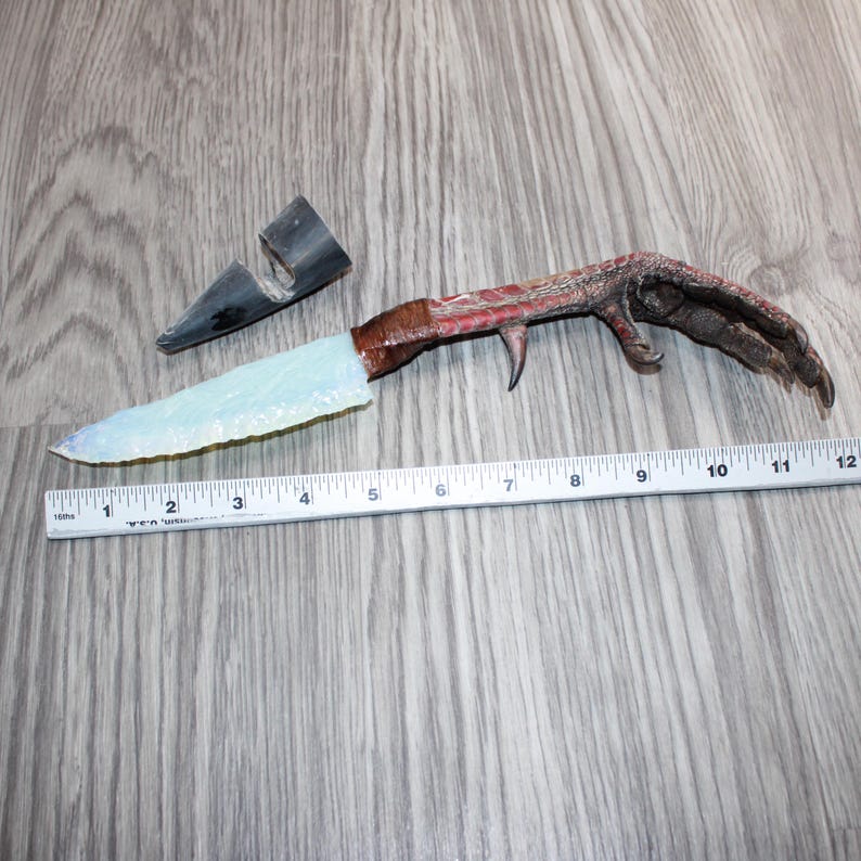 May include: A unique knife with a light blue blade and a handle crafted from a bird's foot. The blade is approximately 12.7 cm long, and the handle is wrapped in brown material. A black sheath is also present.