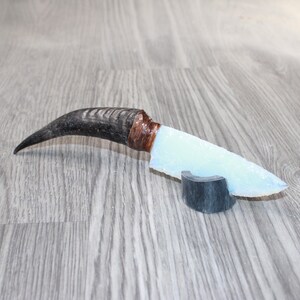 May include: A handcrafted knife with a curved, dark horn handle and a light blue, translucent blade. The handle is wrapped with a copper-colored material. The knife rests on a small, dark gray stand.