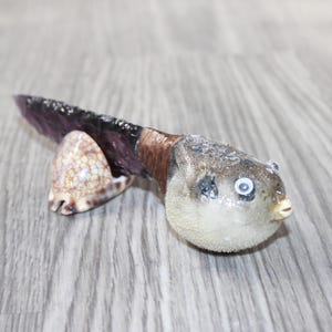 May include: A unique decorative knife featuring a pufferfish handle, a purple blade, and a seashell base. The pufferfish is tan and gray, with white eyes and a small beak. The blade is a dark purple color.