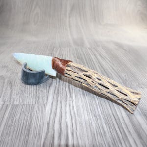 May include: A handcrafted knife with a light blue blade and a textured wooden handle. The handle is wrapped with brown material. The knife rests on a dark gray stand, showcasing its unique design and craftsmanship.