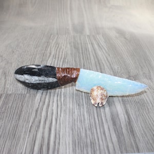 May include: A decorative knife with a black and white stone handle, wrapped in brown material, and a light blue blade. A small, patterned seashell rests near the blade. The knife is displayed on a wooden surface.