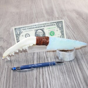 May include: A handcrafted knife with a white jawbone handle, a brown wrapped section, and a light blue blade. A one-dollar bill and a blue pen are next to the knife. A small, curved, white object is also present.