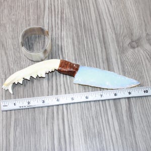 May include: A handmade knife with a light blue blade and a brown handle, next to a white ruler. The blade is approximately 5 inches long. A white bracelet is also in the image.