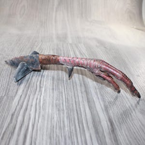 May include: A unique, handmade object resembling a bird's foot, with a blade-like extension. The foot is a deep reddish-brown color, with a textured surface. The blade is dark gray and appears to be made of metal.