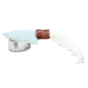 May include: A handcrafted knife with a light blue blade and a brown wrapped handle. The blade is supported by a silver-colored stand, and the handle is connected to a white, curved bone-like structure. The knife is set against a white background.