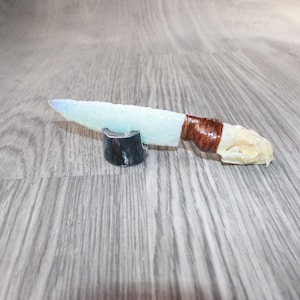 May include: A decorative knife with a light blue blade and a brown wrapped handle. The handle has a white, bone-like end. The knife rests on a small, black and white stand. The blade appears to be made of a translucent material.