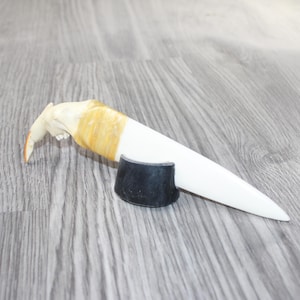 May include: A white, pointed object with a curved, tan end and a yellow band, resting on a black, semi-circular base. The object appears to be a decorative knife or letter opener. The background is a gray wood-grain surface.