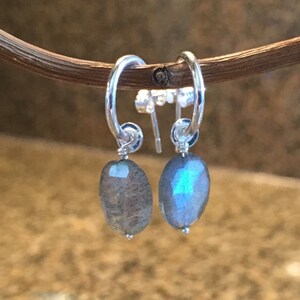 May include: Silver hoop earrings with a dangling oval-shaped labradorite gemstone. The earrings are a light blue color with iridescent shimmer.