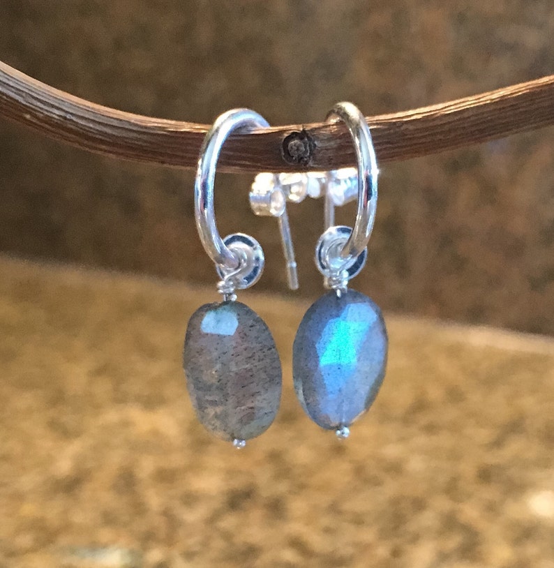 May include: Silver hoop earrings with a dangling oval-shaped labradorite gemstone. The earrings are a light blue color with iridescent shimmer.