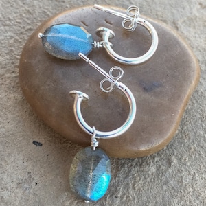 May include: A pair of silver hoop earrings with a dangling oval-shaped labradorite gemstone. The gemstone has a blue-green iridescent sheen.