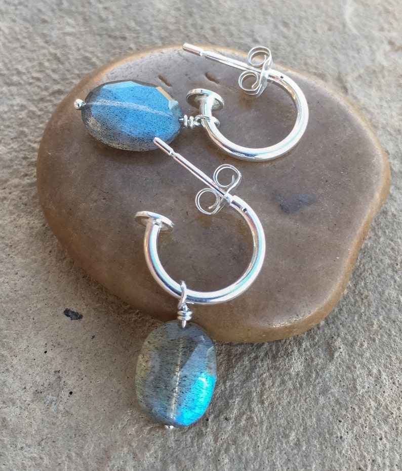 May include: A pair of silver hoop earrings with a dangling oval-shaped labradorite gemstone. The gemstone has a blue-green iridescent sheen.