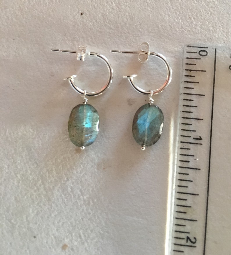 May include: Silver hoop earrings with a dangling oval labradorite gemstone. The earrings are approximately 1 inch long.