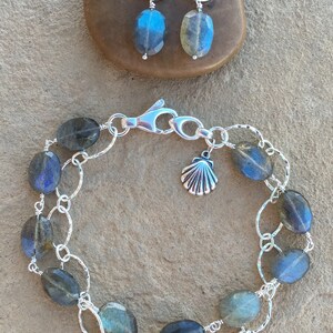 May include: A silver chain bracelet with a seashell charm and oval-shaped labradorite stones. The bracelet is on a light brown textured surface.