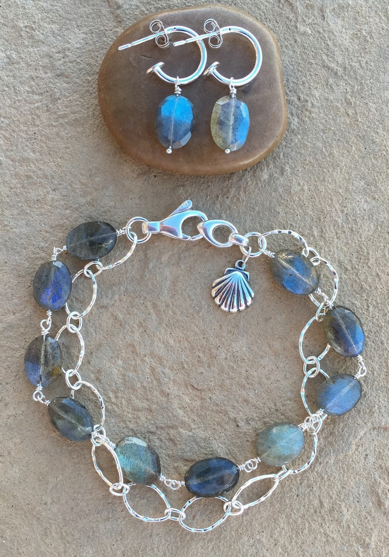 May include: A silver chain bracelet with a seashell charm and oval-shaped labradorite stones. The bracelet is on a light brown textured surface.