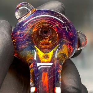 May include: A hand-blown glass pipe featuring a deep purple and blue base with swirling streaks of vibrant orange and yellow. The bowl is circular with a central opening, and the glass surface contains small air bubbles throughout the structure.