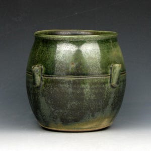 Puede incluir: Una olla de cerámica verde hecha a mano, de forma redondeada, con dos pequeñas asas. La olla tiene una superficie texturizada y un esmalte verde oscuro, con reflejos verdes más claros. Una línea horizontal recorre el centro. Ideal para la decoración del hogar, mide unos 15 cm de alto.