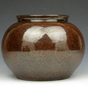 May include: A round, speckled brown ceramic vase with a wide body and a narrow neck. The vase has a glossy finish and a textured surface. The color transitions from a dark brown at the top to a lighter, speckled brown at the base. A decorative piece for the home.